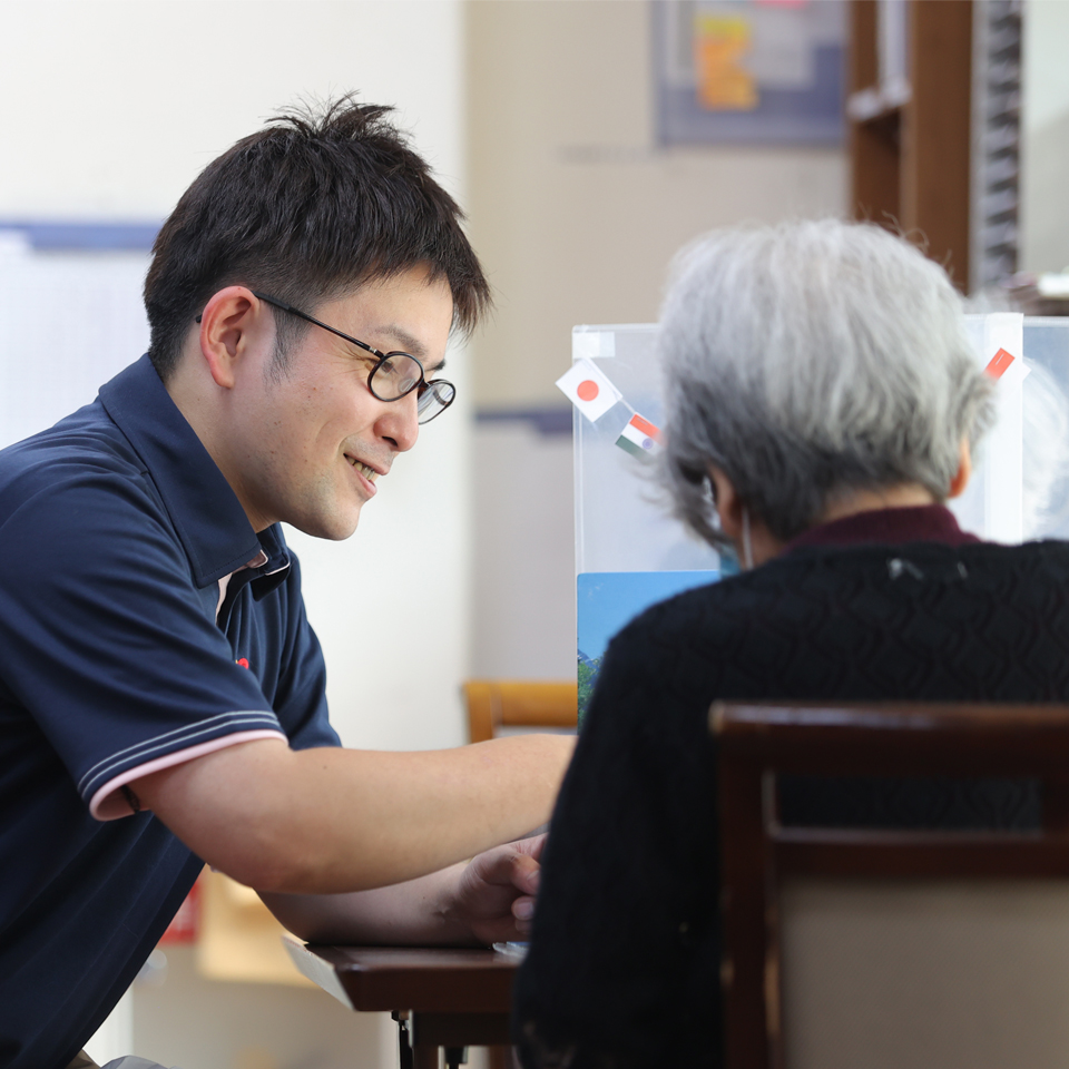 スタッフが高齢者と交流する様子。日本国旗が飾られた室内での交流シーン。